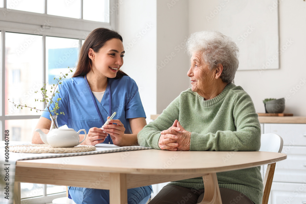 Young caregiver measuring sugar level of senior woman in kitchen