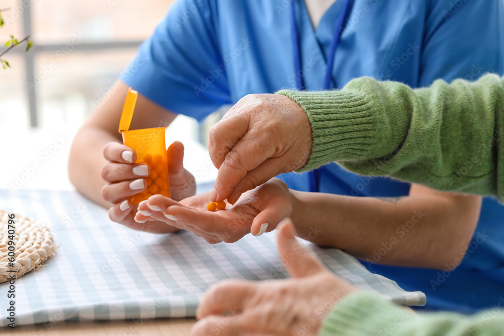 Young caregiver and senior woman taking pills in kitchen, closeup