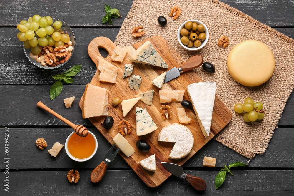 Board with different types of tasty cheese, nuts, honey and grapes on dark wooden background