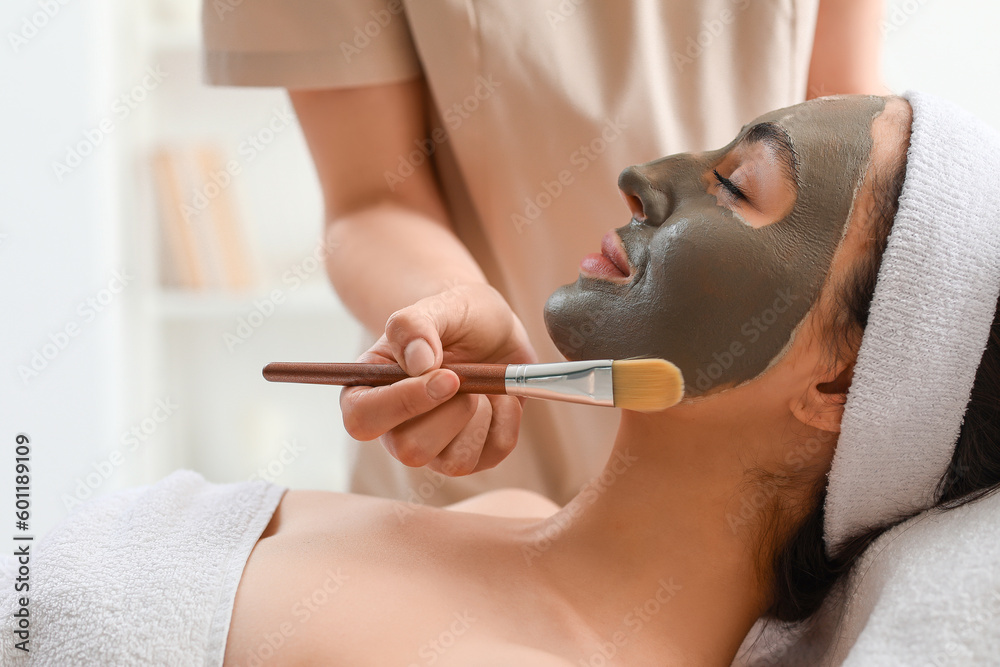 Cosmetologist applying clay mask onto young womans face in salon, closeup