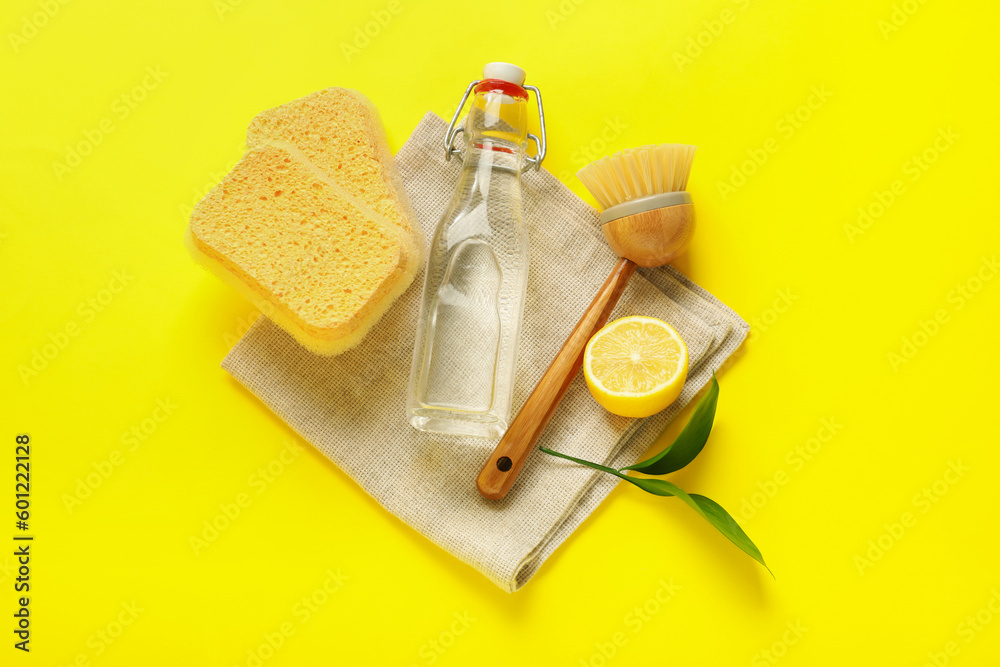 Bottle of vinegar, sponges, brush, napkin and lemon on yellow background