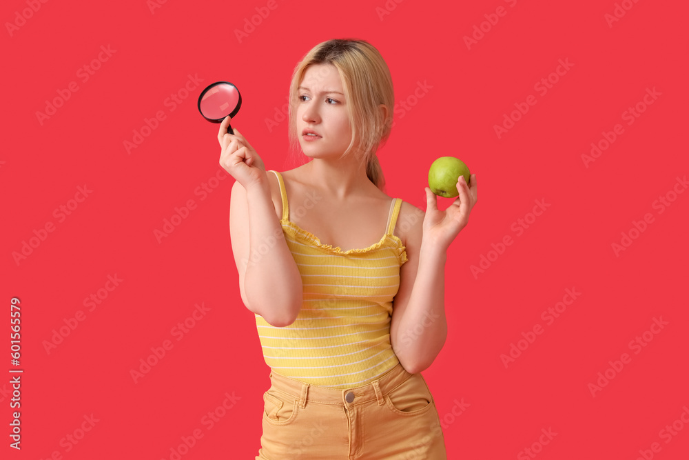 Thoughtful young woman with magnifier and apple on red background