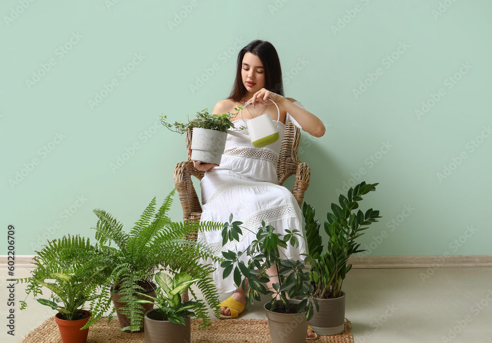 Young woman with watering can and houseplants sitting in armchair near green wall