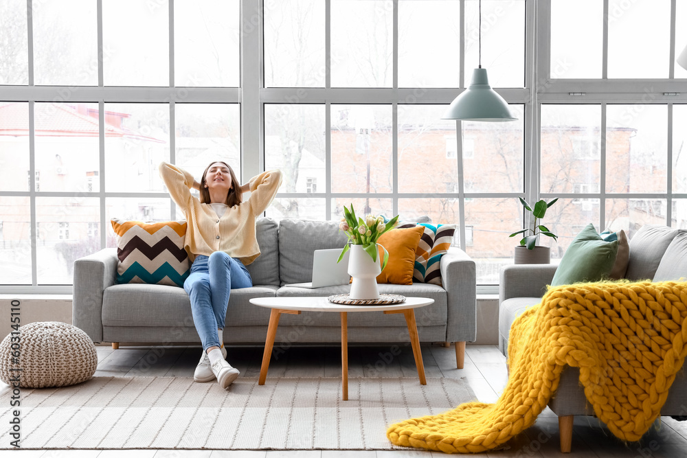 Beautiful young woman sitting on cozy sofa near big window in living room