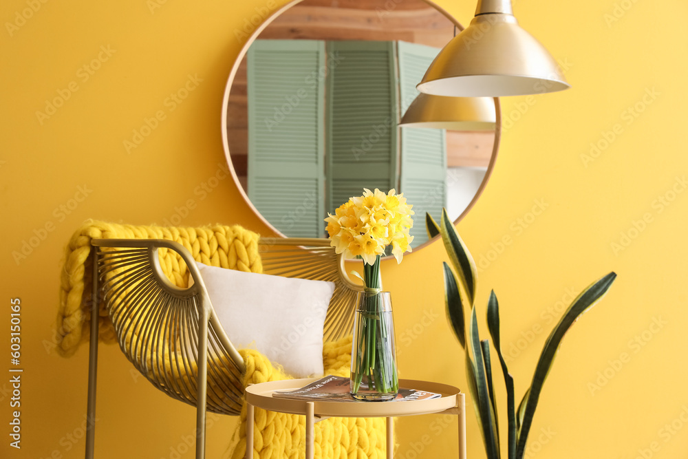 Stylish armchair, vase with blooming narcissus flowers on coffee table and mirror near yellow wall