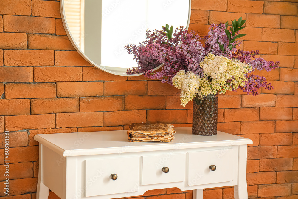 Vase with blooming lilac flowers and old book on table near brown brick wall