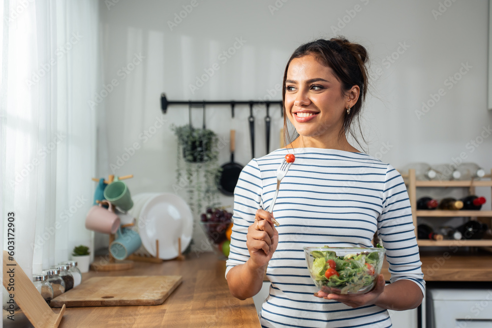 Caucasian young woman eating healthy green salad in kitchen at home.