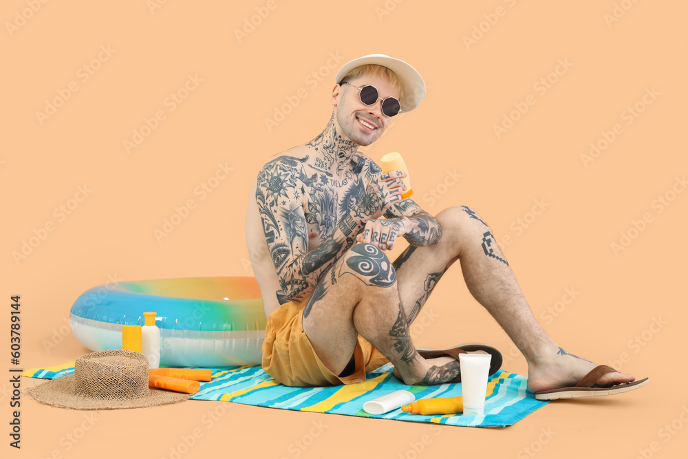 Tattooed man with sunscreen cream and beach accessories on beige background