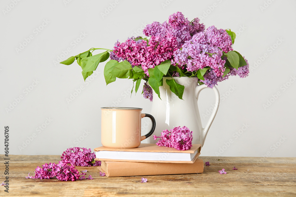 Jug of beautiful lilac flowers with coffee and books on wooden table near white wall