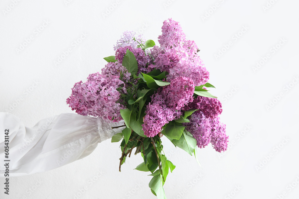 Female hand holding bouquet of beautiful lilac flowers near white wall