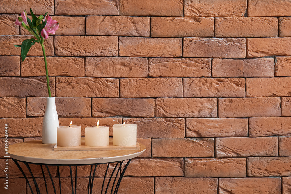 Burning candles and vase with alstroemeria flower on table near brown brick wall