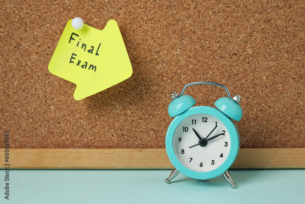 Sticky note with text FINAL EXAM hanging at cork board and alarm clock on blue table