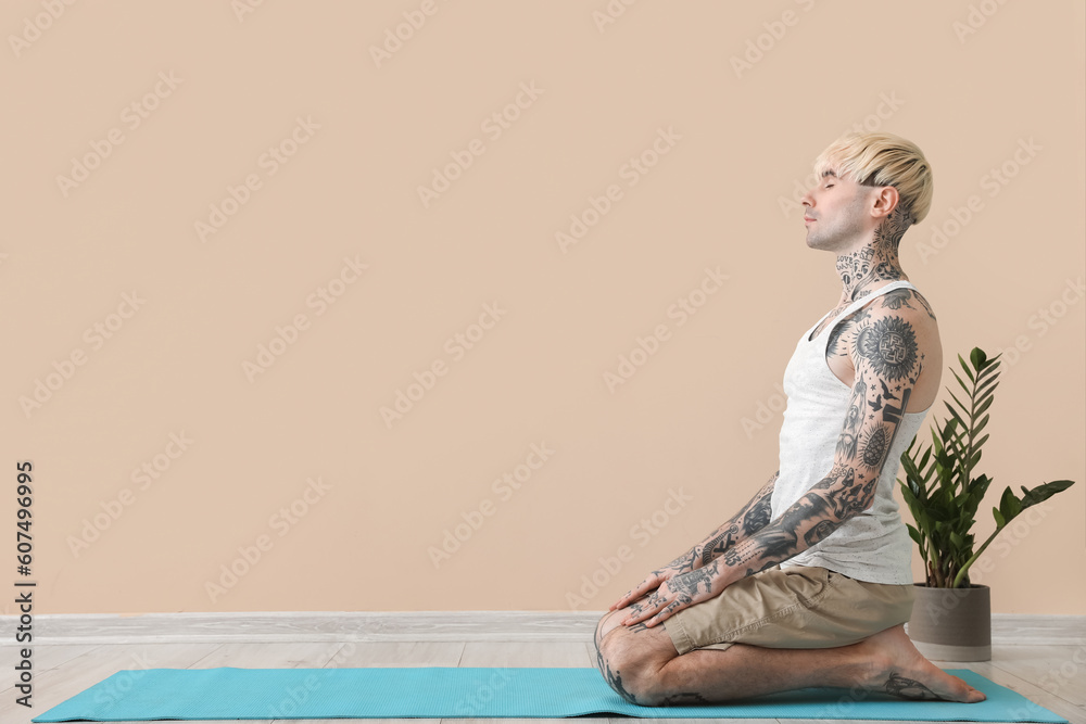 Tattooed young man meditating near beige wall