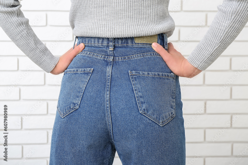 Young woman in stylish jeans near light brick wall, back view