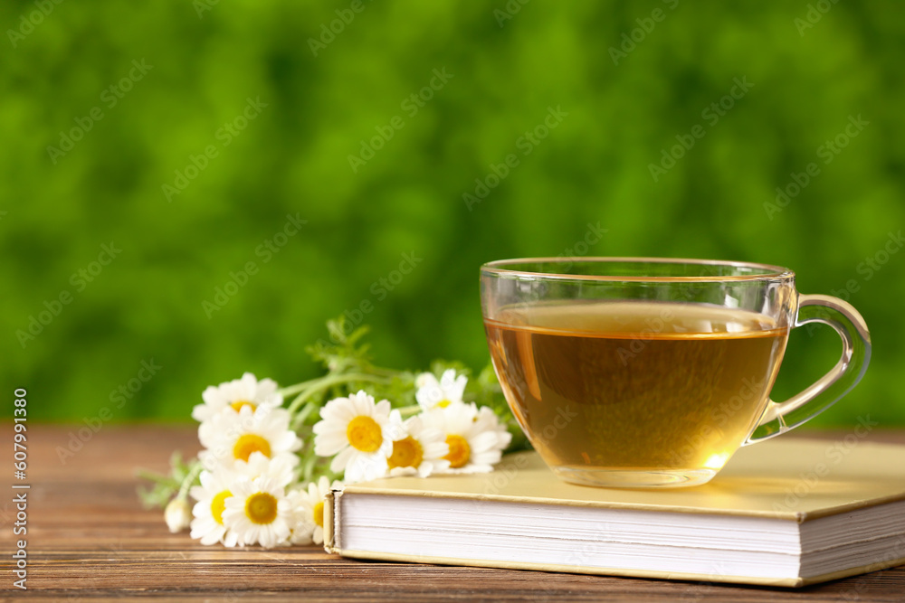 Book and cup of tea with beautiful chamomile flowers on wooden table outdoors