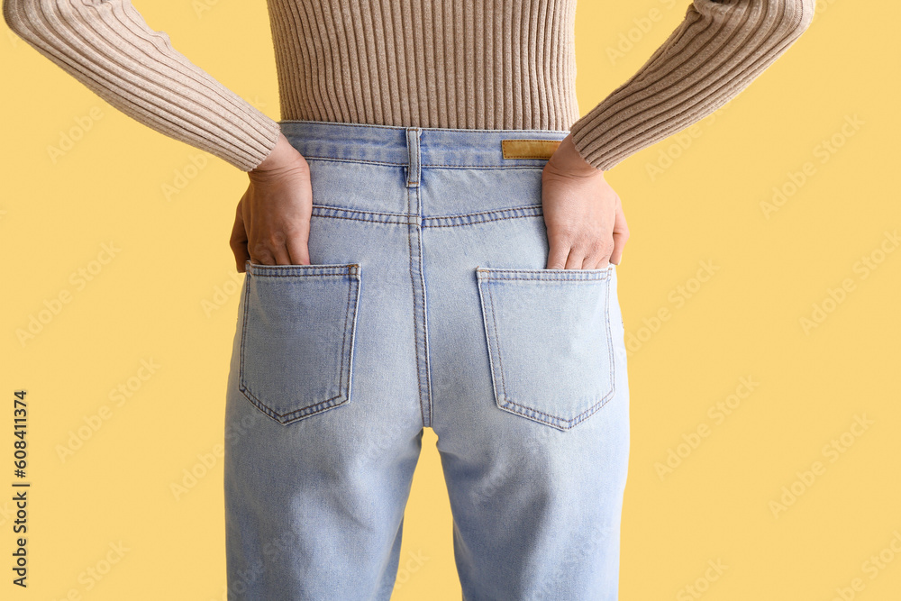 Young woman in stylish jeans on yellow background, back view