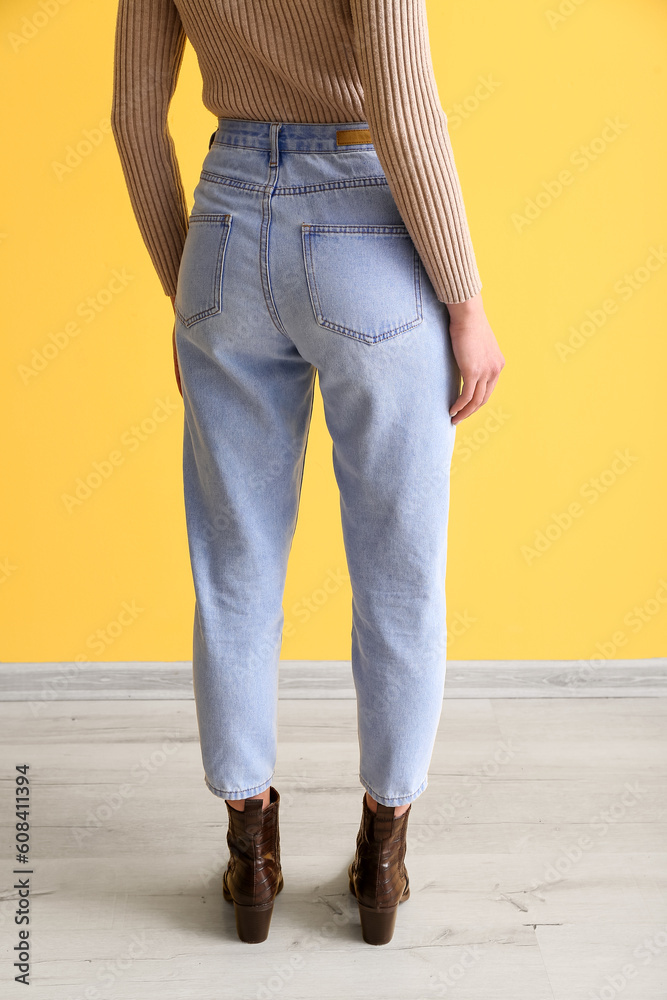 Young woman in stylish jeans near yellow wall, back view