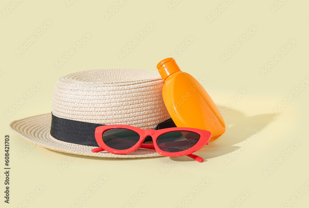 Bottle of sunscreen cream with sunglasses and summer hat on pale yellow background