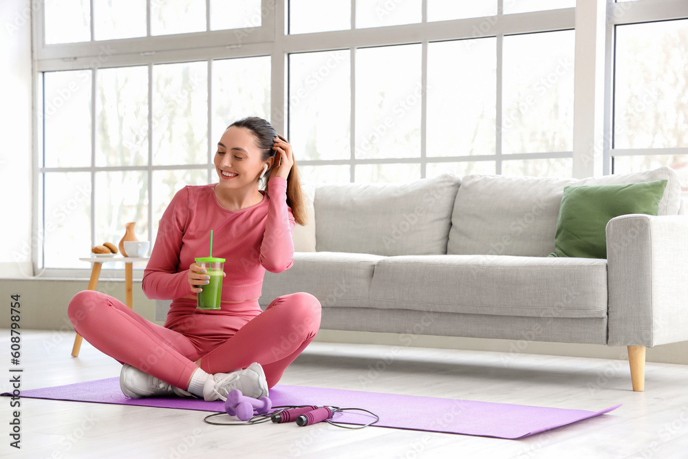 Sporty young woman in earphones with glass of vegetable juice at home