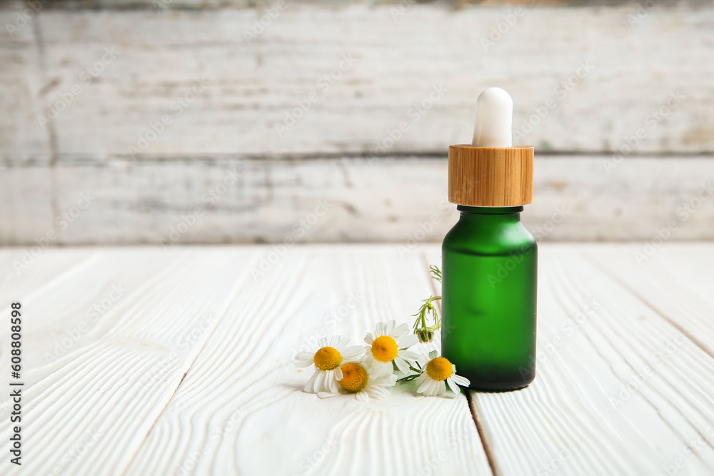 Bottle of essential oil and chamomile flowers on light wooden table