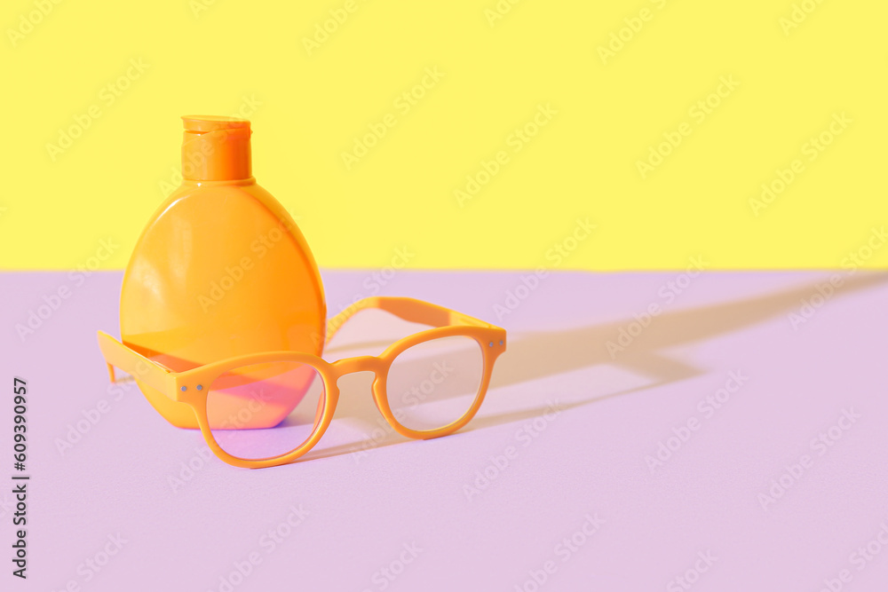 Bottle of sunscreen cream with sunglasses on lilac table near yellow wall