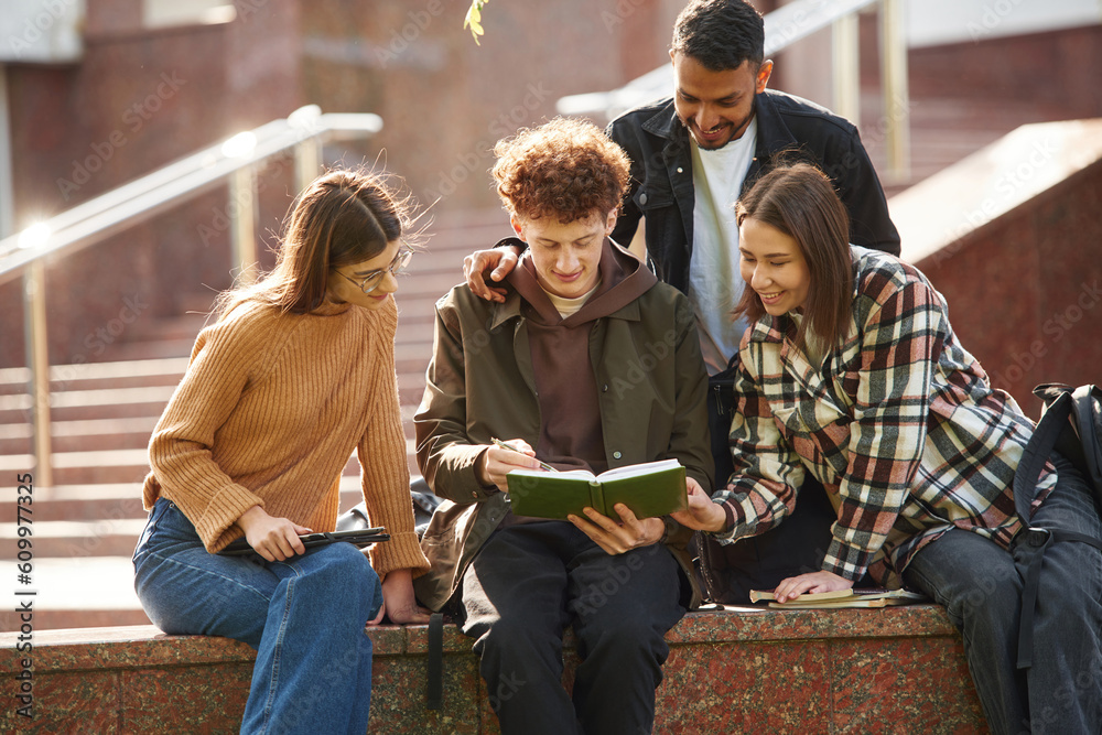 Sitting, reading educational material. Four young students in casual clothes are together outdoors