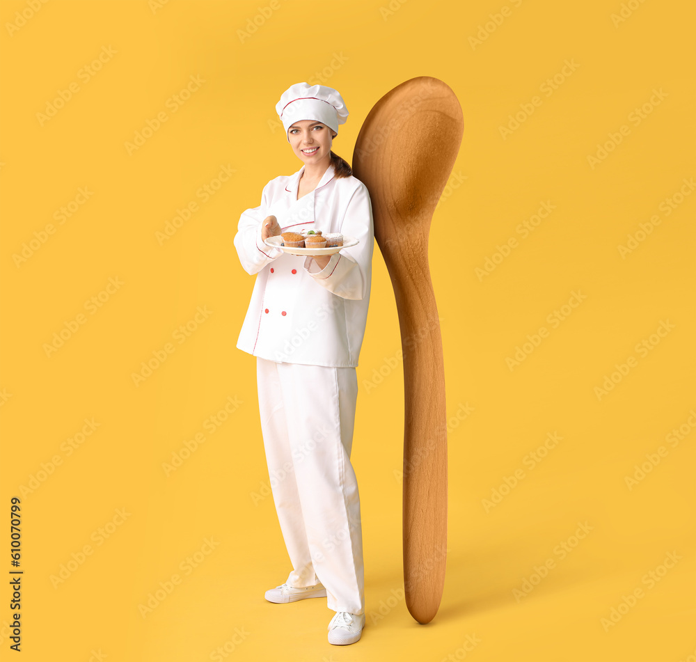 Female confectioner with tasty muffins and big wooden spoon on yellow background