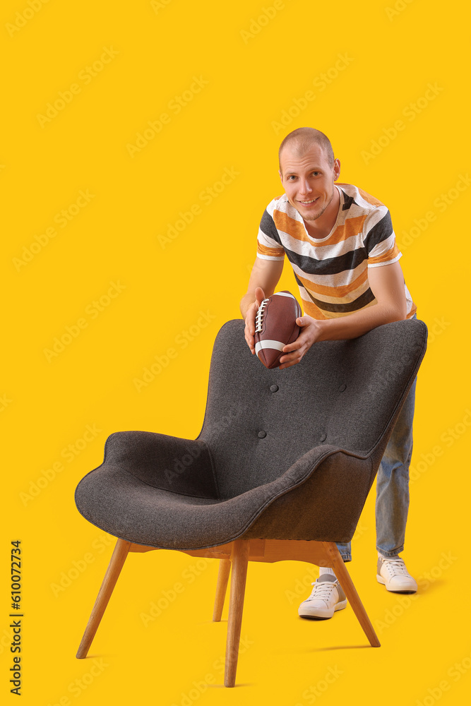 Handsome man with rugby ball beside black armchair on yellow background