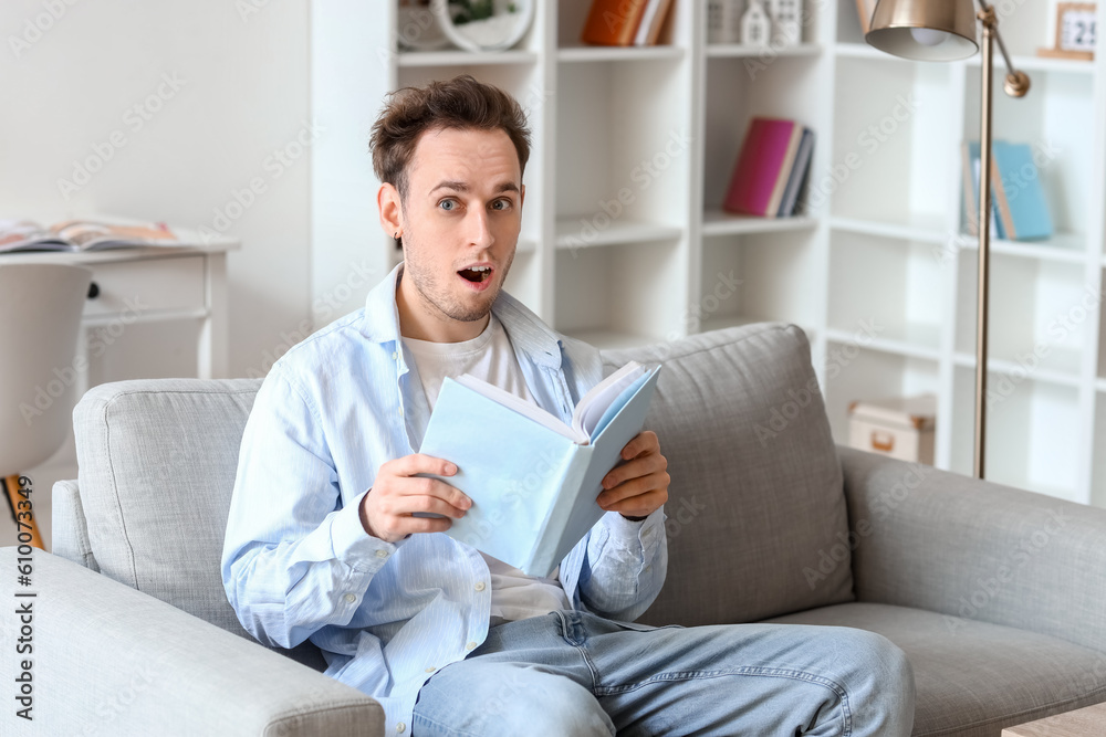 Shocked young man reading book on sofa at home