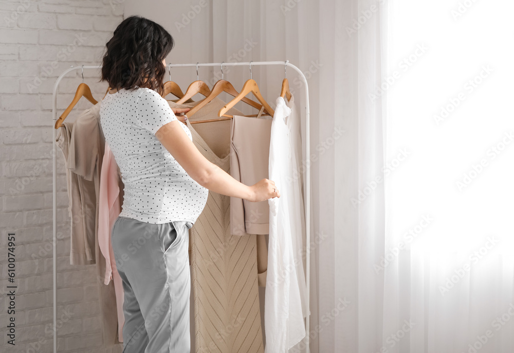 Young pregnant woman near rack with clothes in bedroom