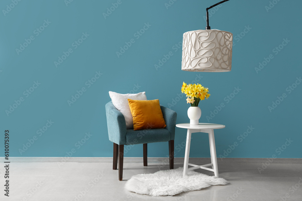 Cozy armchair with cushions and blooming narcissus flowers on coffee table near blue wall