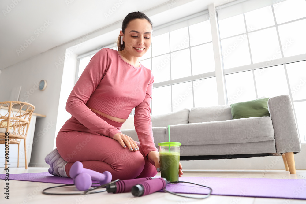 Sporty young woman in earphones with glass of vegetable juice at home