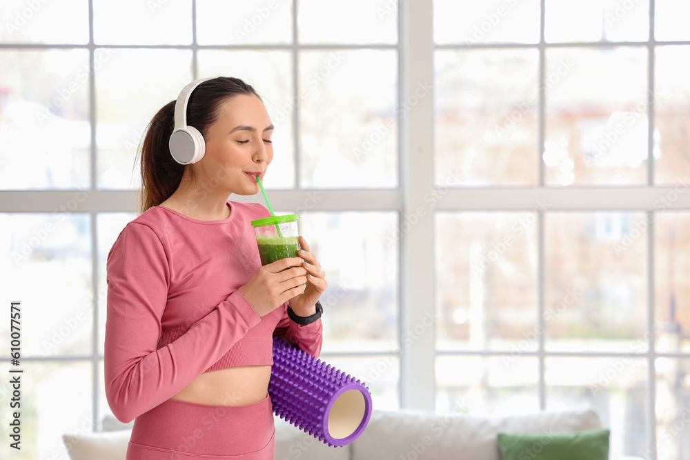 Sporty young woman in headphones drinking vegetable juice at home