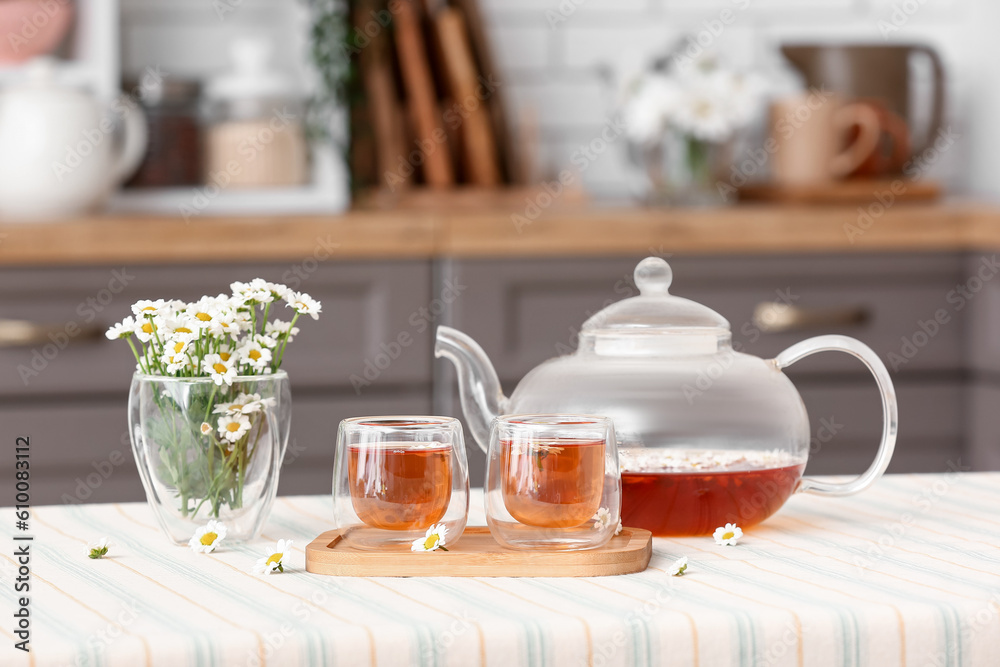 Wooden tray with teapot, cups of natural chamomile tea and flowers on table in kitchen