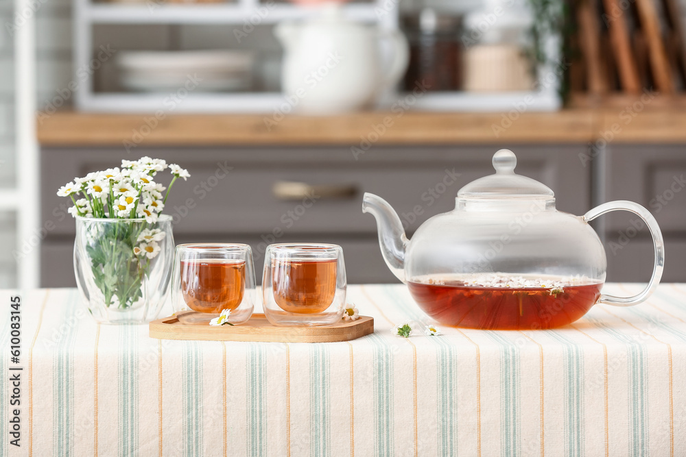 Wooden tray with teapot, cups of natural chamomile tea and flowers on table in kitchen