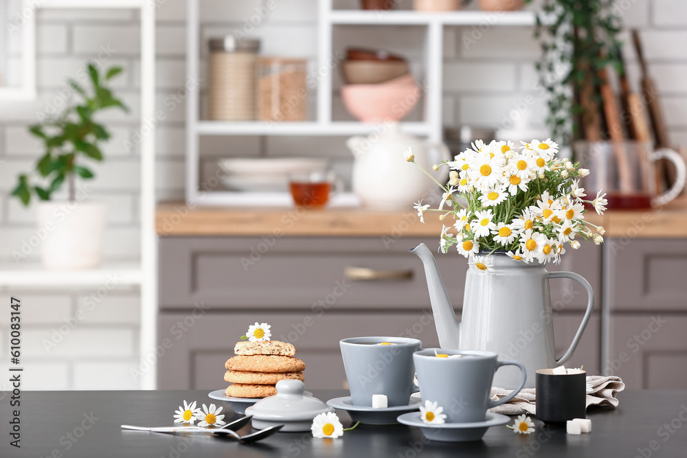 Watering can with flowers, cups of natural chamomile tea and cookies on black table in kitchen