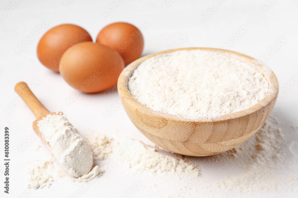 Wooden bowl with wheat flour, scoop and eggs on white background