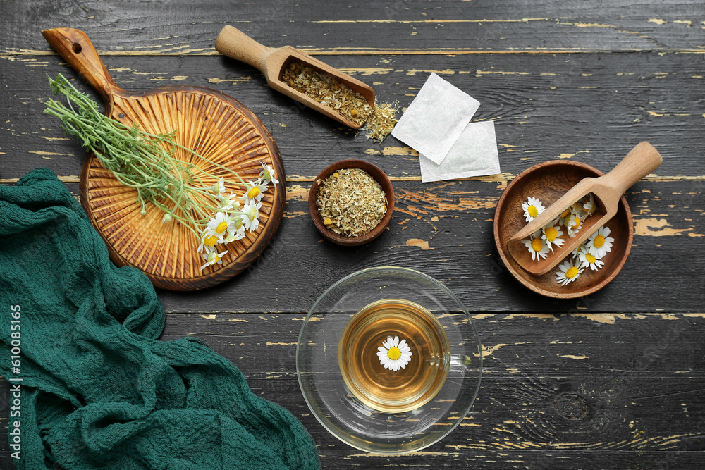 Cup of hot tea and bowl with dried chamomile on black wooden background