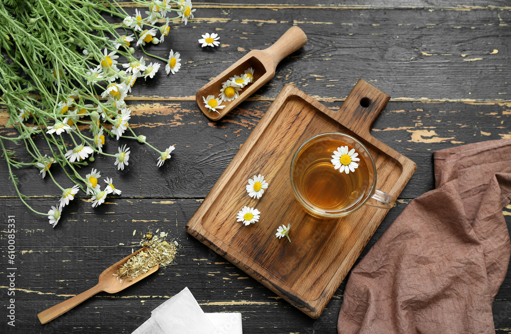 Board with cup of hot chamomile tea and scoops on black wooden background