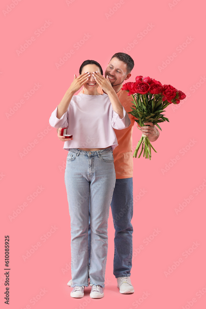 Young man with engagement ring and flowers closing his girlfriends eyes on pink background