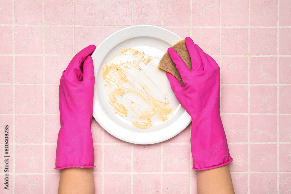 Female hands in rubber gloves washing dirty plate on pink tile background