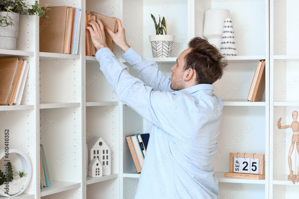 Young man taking book from shelf at home