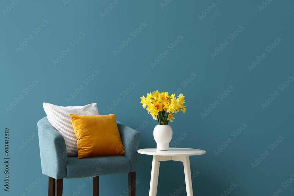 Cozy armchair with cushions and blooming narcissus flowers on coffee table near blue wall