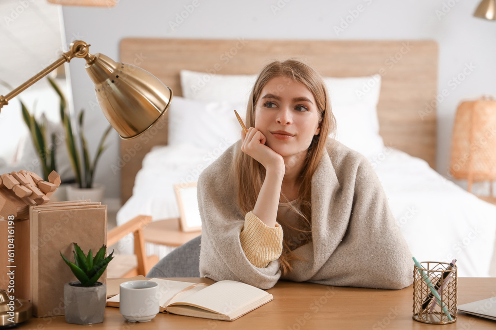 Dreaming young woman with notebook at table in bedroom