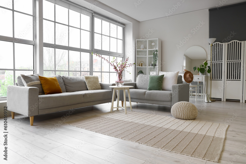 Interior of living room with grey sofas and blooming sakura branches on coffee table near big window