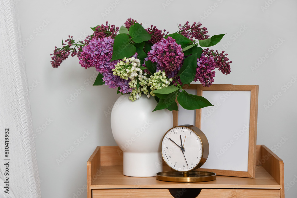 Vase with lilac flowers, clock and frame on table in bedroom