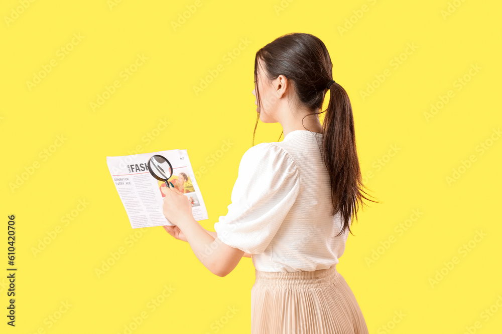 Young woman with magnifier reading newspaper on yellow background