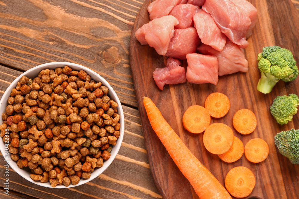 Bowl with dry pet food, raw meat and natural products on wooden background