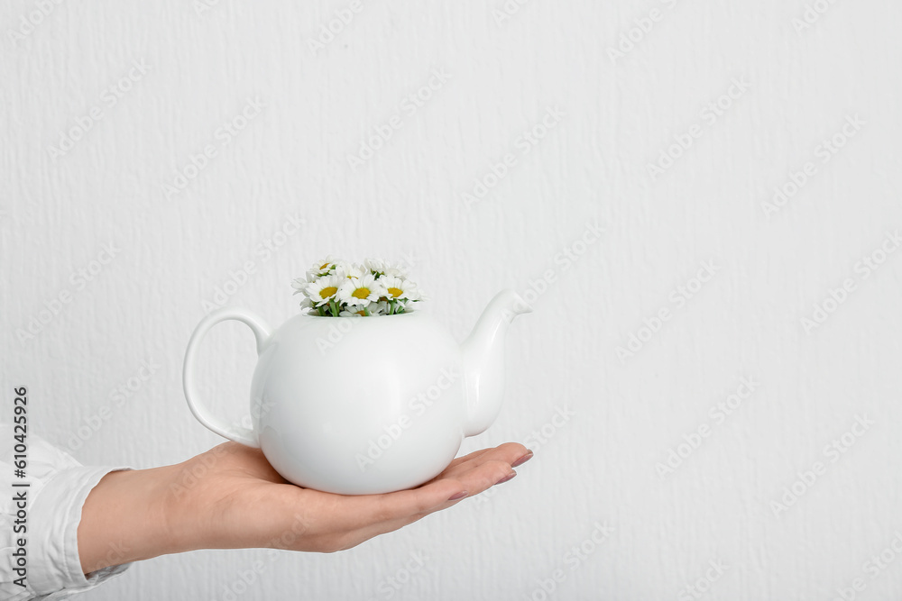 Female hand with teapot of chamomile tea and flowers near white wall