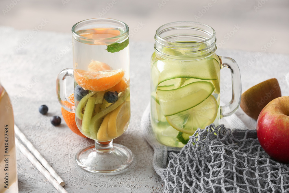 Glass and mason jar of infused water with different sliced fruits on grey table